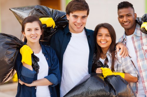 Four young volunteers, two women and two men, standing outdoors during daytime, each holding large black trash bags filled with rubbish. The individuals are smiling and appear to be participating in a community rubbish removal activity, wearing casual clothing and yellow gloves for handling waste. The background shows a residential setting with blurred greenery and houses, suggesting a neighbourhood clean-up effort. The scene captures the team’s positive spirit and their engagement in rubbish clearance, aligning with services offered by House Clearance Redhill for local property and waste management in the Redhill area. The natural lighting highlights the textures of the filled rubbish bags, the casual outfits, and the friendly expressions of the volunteers, emphasizing their contribution to environmental cleanliness and community improvement.