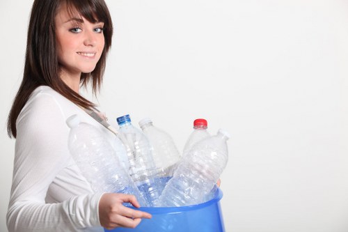 A young woman with shoulder-length brown hair and a light complexion is standing against a plain white background, smiling gently as she holds a blue plastic bin filled with several transparent plastic bottles. The bottles vary in size and shape, some with blue caps, others with red or white caps, and appear to be empty, used for beverages or water. She is wearing a long-sleeved white top, and her left hand supports the bottom of the bin while her right hand grips its side. The scene gives a clean and organized impression, with the focus on her handling the bottles that are associated with waste that may be collected and recycled by a rubbish removal service such as House Clearance Redhill. The neutral setting emphasizes the action of waste disposal in a domestic context, subtly connecting to local rubbish clearance efforts in the Redhill area and surrounding towns with postcode RH.