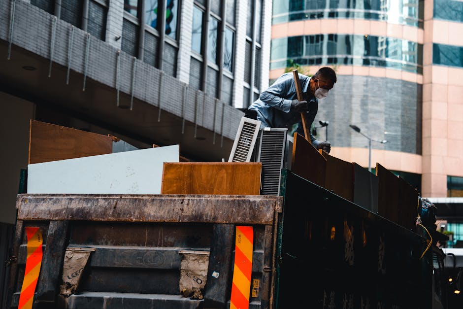 A worker wearing a white shirt, dark trousers, gloves, and a face mask is standing on the edge of a large open-top waste truck, which is parked outdoors in an urban area with modern glass and concrete