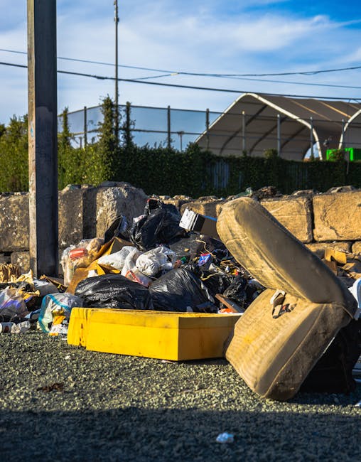 In the foreground, a pile of mixed household waste, including brown cardboard boxes, plastic bags, and dark-colored refuse bags, is situated on a paved driveway adjacent to a residential building. The