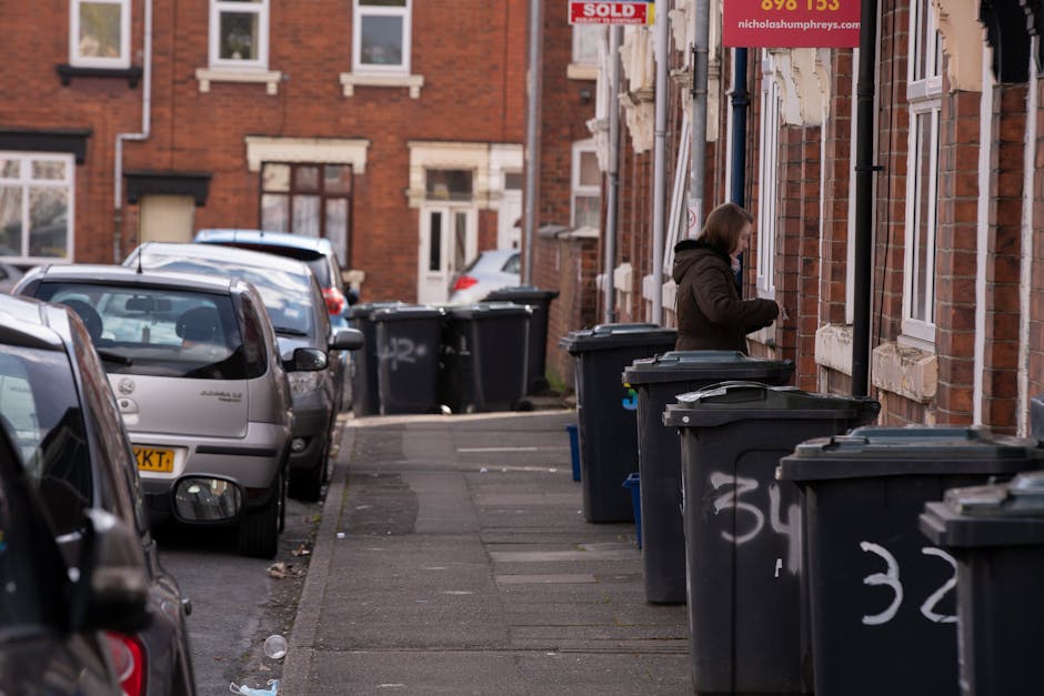 A large outdoor scene showing a significant accumulation of waste and household rubbish piled next to a sidewalk in an urban setting. The debris includes various materials such as cardboard boxes, pla