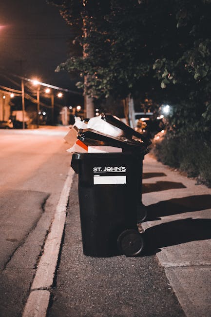A middle-aged man with dark hair, wearing a black T-shirt with white text on the front, is seen emptying a public recycling or waste bin on a paved walkway. He is leaning forward, holding a large whit
