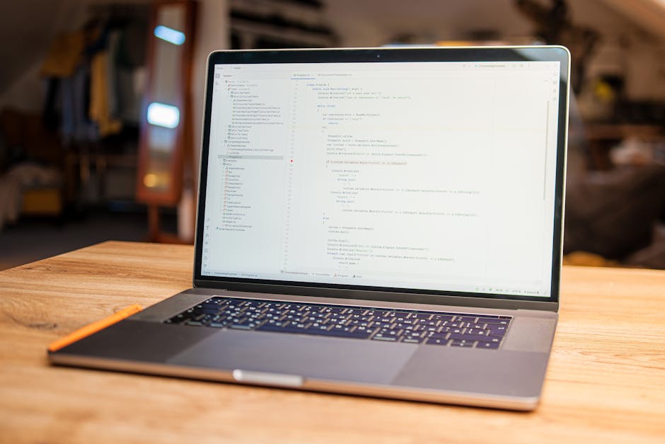 A close-up view of a workspace featuring an open silver laptop with a black keyboard, displaying lines of colorful computer code on the screen. To the left of the laptop, there is a spiral-bound noteb