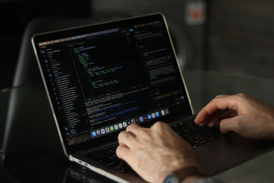 The image shows a person seated at a desk in a modern, well-lit office environment, working on a computer with a large monitor displaying lines of colorful code in a text editor. The individual, seen 