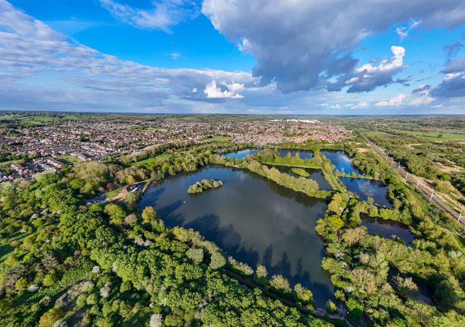 An aerial view of a large urban park with several interconnected lakes, surrounded by dense greenery and trees with bright green foliage. The park is bordered by a residential area with numerous house